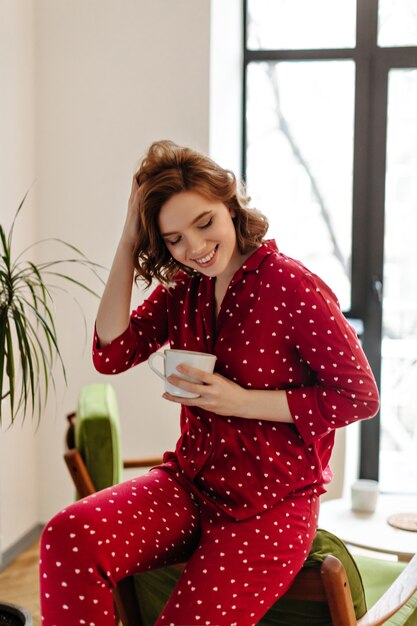 Pretty woman in red pajama sitting on armchair and touching curly hair. Indoor shot of laughing young woman with cup of coffee.