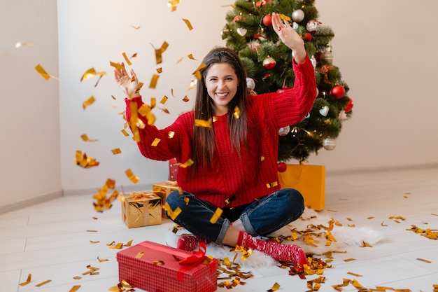 선물 및 선물 상자에 둘러싸인 황금 색종이를 던지고 크리스마스 트리에서 집에 앉아 빨간 스웨터에 예쁜 여자