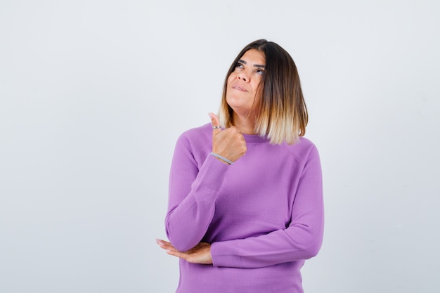 Pretty woman showing thumb up, looking upward in purple sweater and looking dreamy , front view.