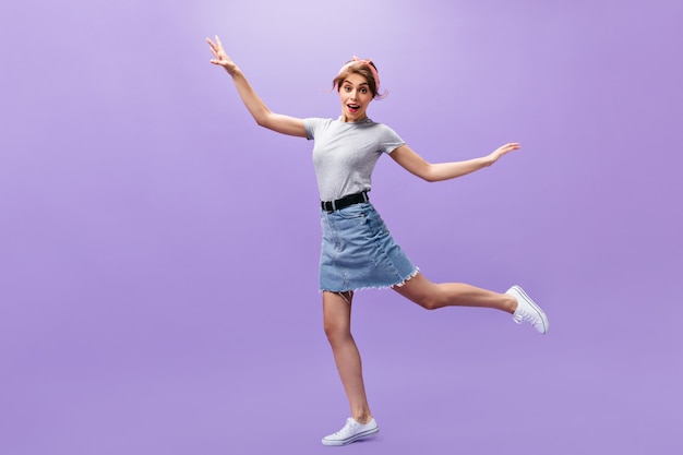 Красивая женщина в юбке и рубашке прыгает на фиолетовом фоне. Смешная маленькая девочка со стильной прической в белых кроссовках позирует.