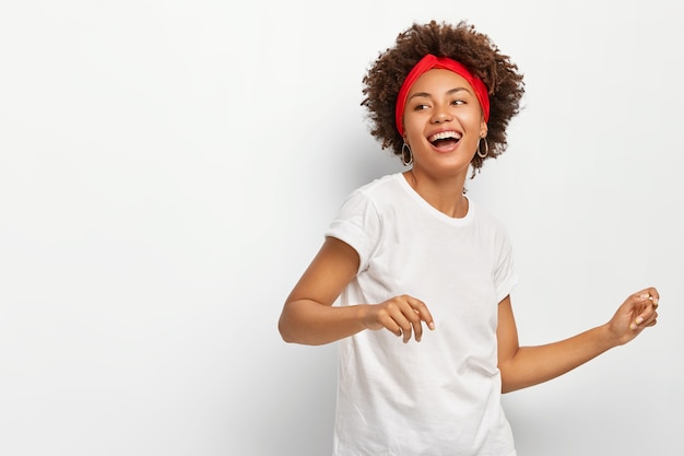 Pretty woman stands sideways, feels energized dances actively to music wears red headband casual white t shirt poses indoor