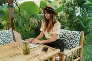 Free photo pretty woman in straw hat using lap top while traveling in asia  working remotely in stylish tropical cafe