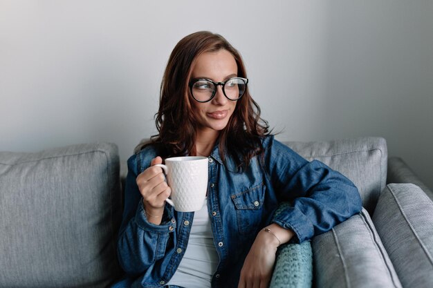 Pretty woman student is wearing glasses and denim shirt is drinking morning coffee and looking at window before working day