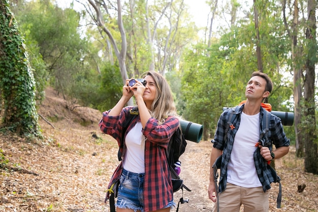 Pretty woman taking photo of landscape with camera and traveling with boyfriend. Caucasian tourists hiking together. Male backpacker looking at scenery. Tourism, adventure and summer vacation concept
