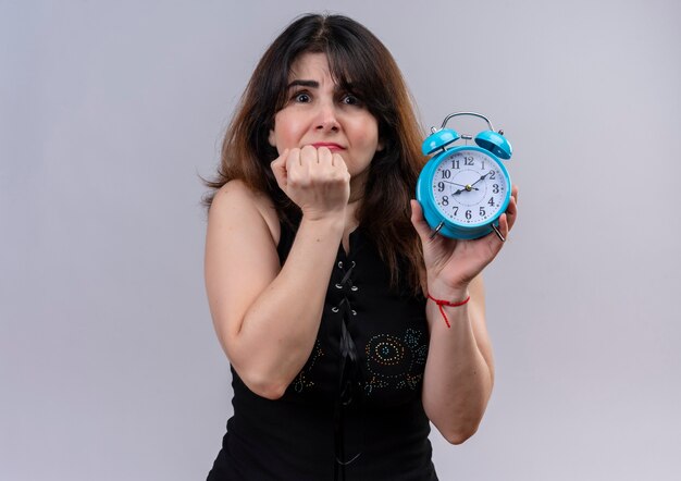 Pretty woman wearing black blouse holding clock scared of being late over gray background