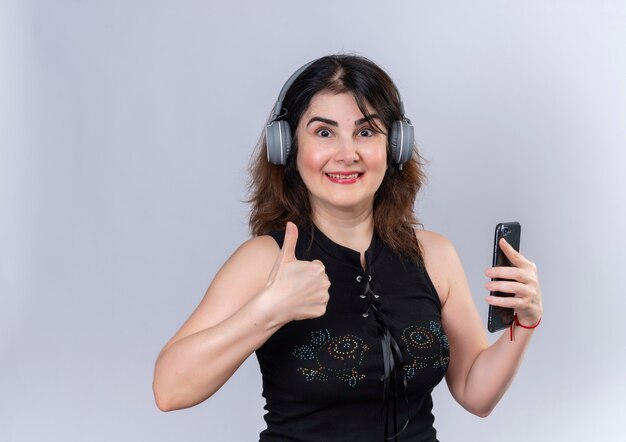 Pretty woman wearing black blouse looking  doing happy thumbs up in headphones holding phone