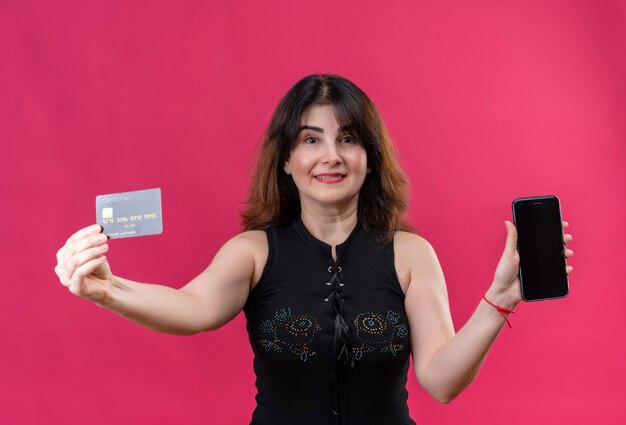 Pretty woman wearing black blouse looking  happily holding credit card and phone