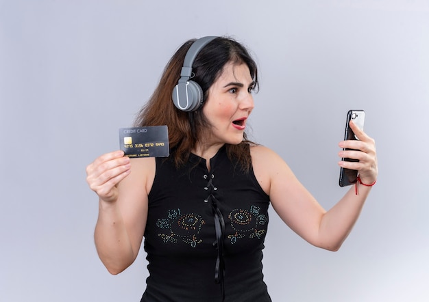 Bella donna che indossa camicetta nera, parlando al telefono felicemente sorpreso in possesso di una carta di credito