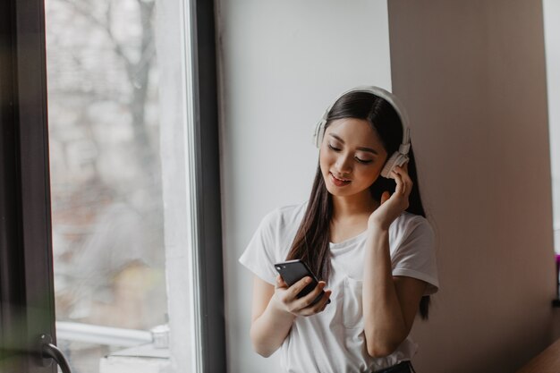 Pretty woman wearing white T-shirt, holding phone and listening to music on headphones by window