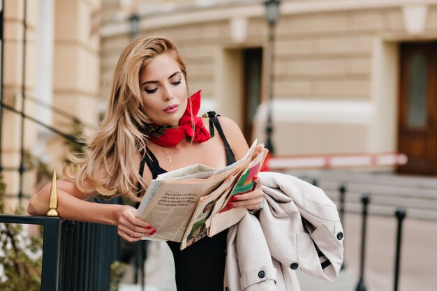 Pretty woman wears golden pendant reading newspaper while waiting friend on the street