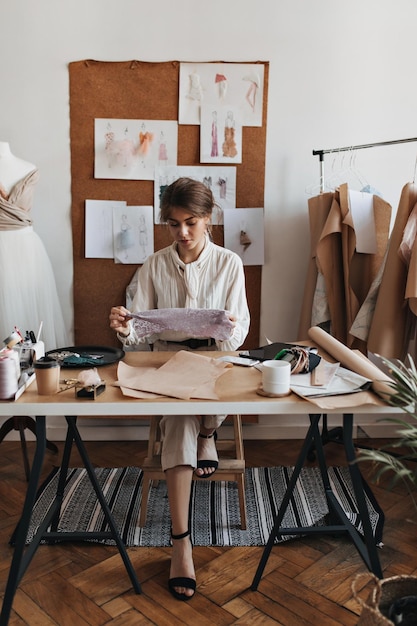 Pretty woman in white blouse looks at lace table Brunette lady in beige shirt and pants posing in designer office