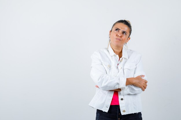 Pretty woman in white jacket keeping arms folded, looking up and looking thoughtful , front view.