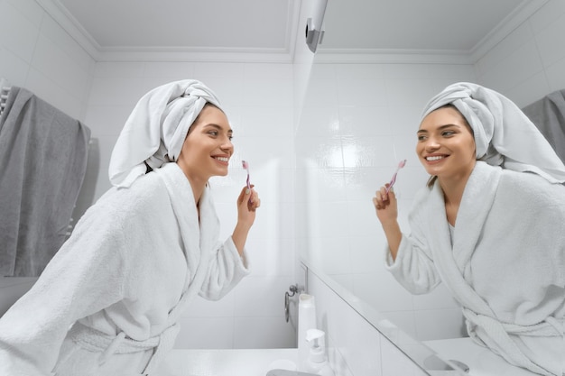 Pretty woman in white robe with towel on head brush teeth in bathroom