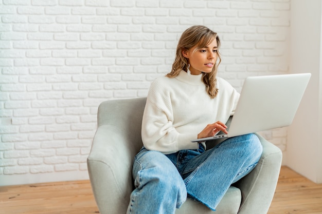 Красивая женщина со светлыми волнистыми волосами, сидя на диване у себя дома, работает на портативном компьютере