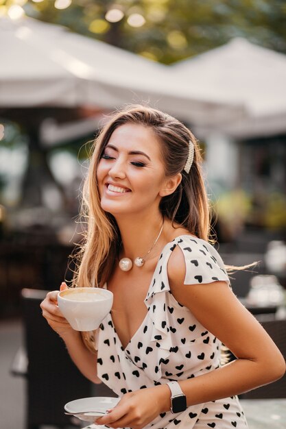 Красивая женщина с откровенной улыбкой смеется, сидя в летнем кафе с чашкой капучино