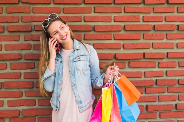 Pretty woman with colourful shopping bags talking by phone at brick wall 