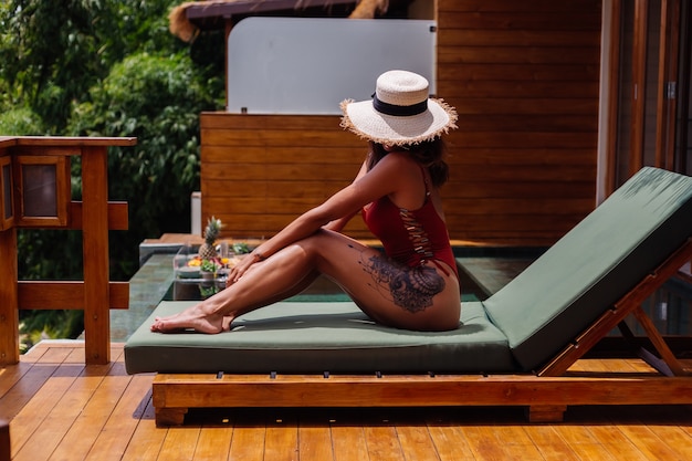 Pretty woman with fit perfect tanned body bronze skin lies on green sunbed at luxury tropical villa in one piece swimsuit with straw hat cover face.