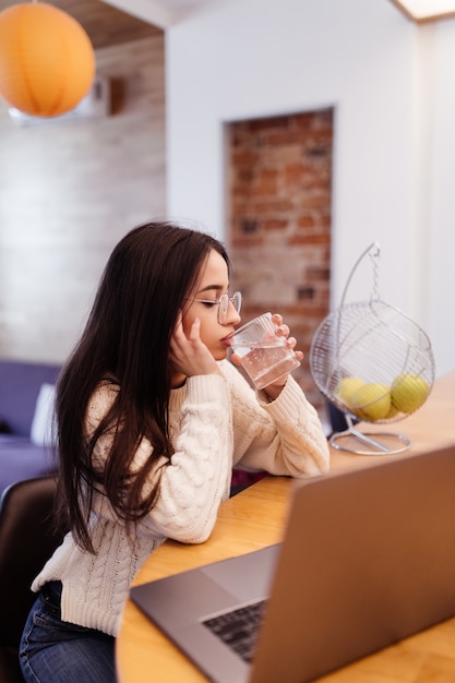 Pretty woman with lond black hair is working on her laptop and drinking water on the kitchen