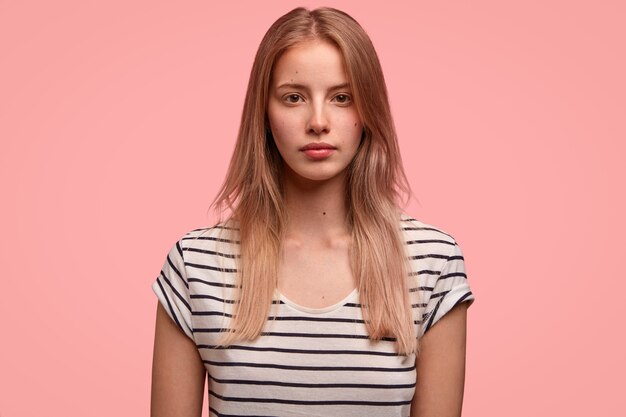 pretty woman with serious expression, healthy skin, light hair, dressed in striped t shirt, poses over pink wall has appealing appearance