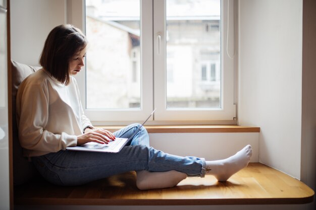 Pretty woman with short hair is working on laptop while sitting on wide windowhill in daily time