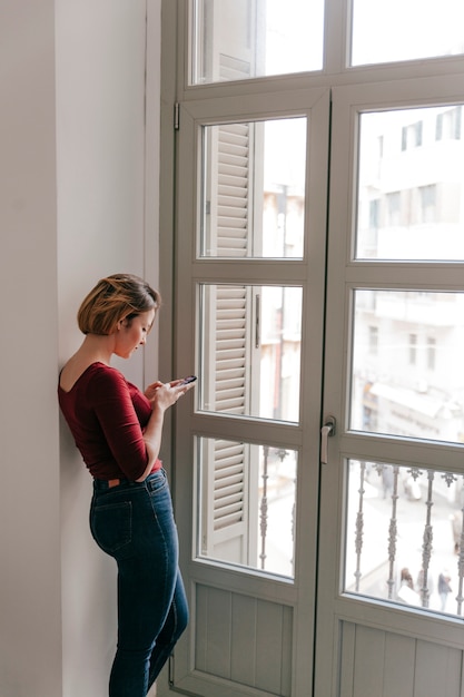 Foto gratuita donna graziosa con lo smartphone vicino alla finestra