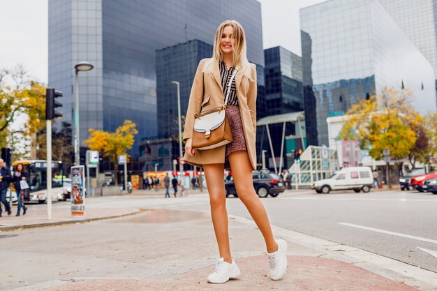 通りを歩いて驚きの顔を持つきれいな女性。ベージュのコートとスニーカーを着用。ニューヨーク。完璧な長い脚。エレガントな外観。