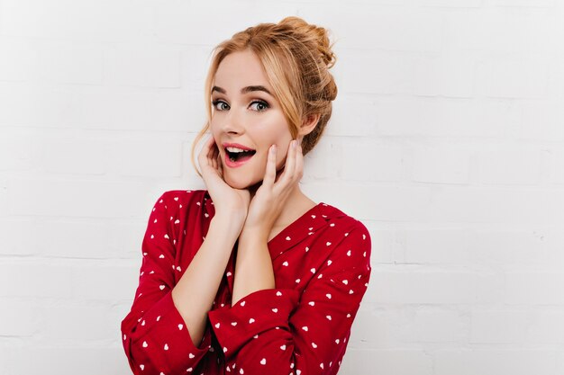 Pretty woman with surprised smile touching her face on white wall. European fair-haired girl posing in red pajamas.