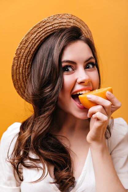 Pretty woman with wavy hair bites juicy orange and looks into camera against isolated background.