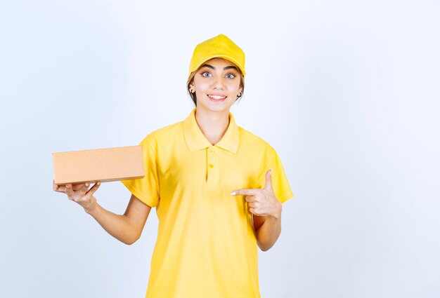 A pretty woman in yellow uniform pointing at a brown blank craft paper box .