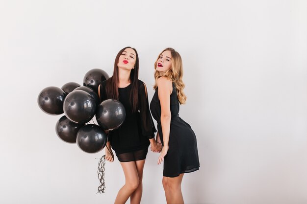 Pretty women in similar dresses holding hands and posing with kissing face expression at party