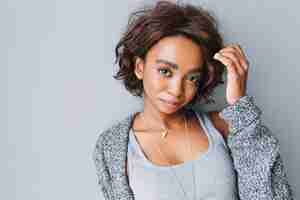 Free photo pretty young african girl with short curly hairstyle on gray wall. wearing gray cardigan, knitted jacket, shirt, necklace with triangle and stylish earrings.