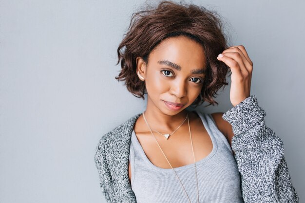 Pretty young african girl with short curly hairstyle on gray wall. Wearing gray cardigan, knitted jacket, shirt, necklace with triangle and stylish earrings.