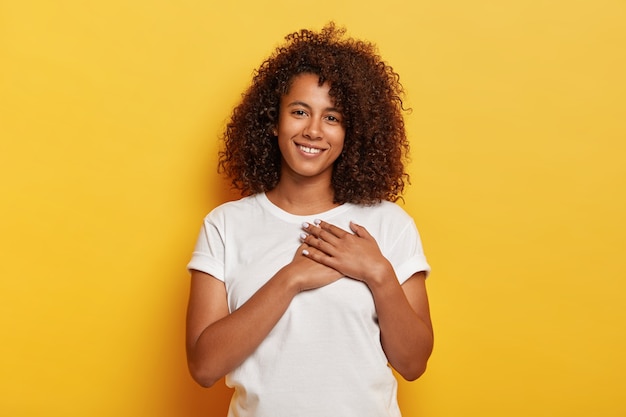 Pretty young Afro American female keeps palms near heart, expresses gratitude