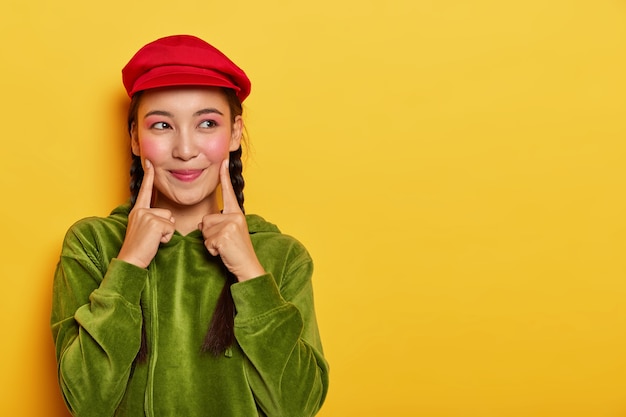Симпатичная молодая азиатская модель держит оба указательных пальца на щеках, смотрит в сторону с мечтательным выражением лица, у нее минимальный макияж, в красном берете и зеленой бархатной толстовке.