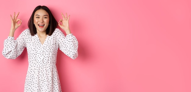 Free photo pretty young asian woman in dress showing okay sign praising and showing approval looking satisfied