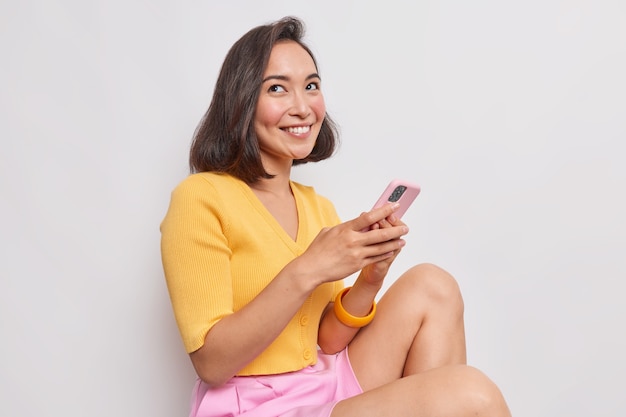 Pretty young Asian woman with bob hairstyle smiles gently has dreamy expression sits against white wall holds mobile phone sends text messages wears yelow jumper pink shorts feels happy