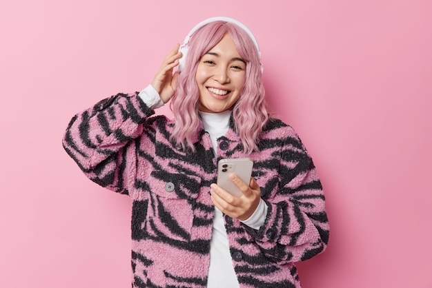 Pretty young asian woman with dyed hair listens music via stereo headphones holds mobile phone dressed in winter jacket has good mood isolated over pink background. people and hobby concept