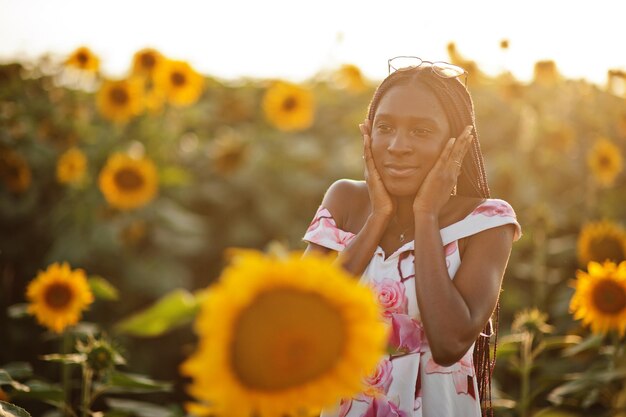 Красивая молодая чернокожая женщина в летнем платье позирует на поле подсолнухов