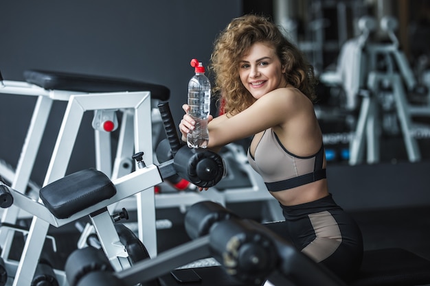 Довольно молодая блондинка спортсменка расслабляется после тренировки и держит бутылку с водой в тренажерном зале