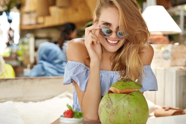 Pretty young blonde woman looks through sunglasses, poses at camera with positive expression, surrounded with exotic cocktail, spends leisure time in exotic cafe, tastes local dish and drinks