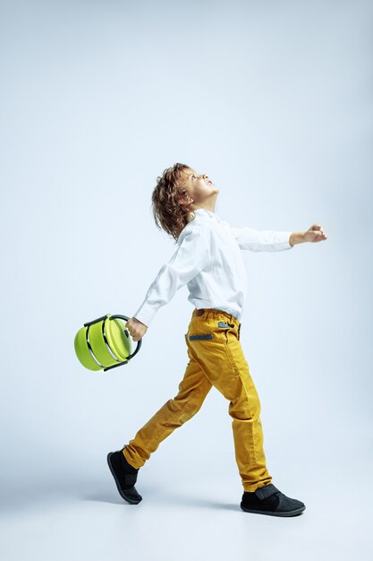 Pretty young boy in casual clothes on white wall. Caucasian male preschooler with bright facial emotions holding lunch bag. Childhood, expression, having fun. Dreamful going and jumping.