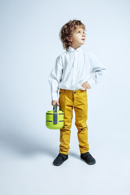 Pretty young boy in casual clothes on white  wall. Caucasian male preschooler with bright facial emotions holding lunch bag. Childhood, expression, having fun. Looks serious, dreamful.