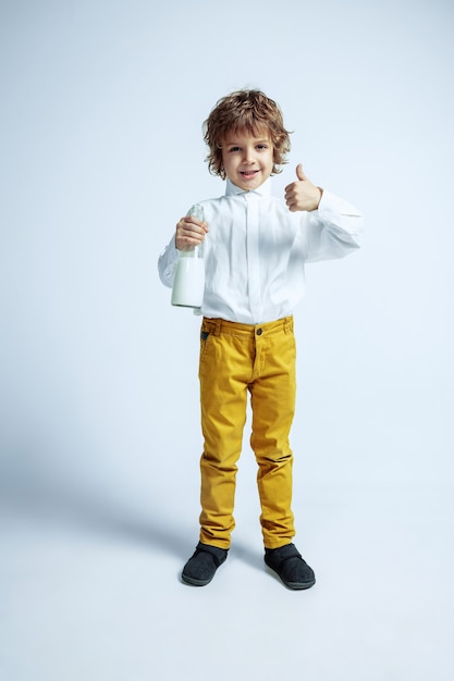 Pretty young boy in casual clothes on white wall. Fashionable posing. Caucasian male preschooler with bright facial emotions. Childhood, expression, having fun. Drinking milk, thumb up.