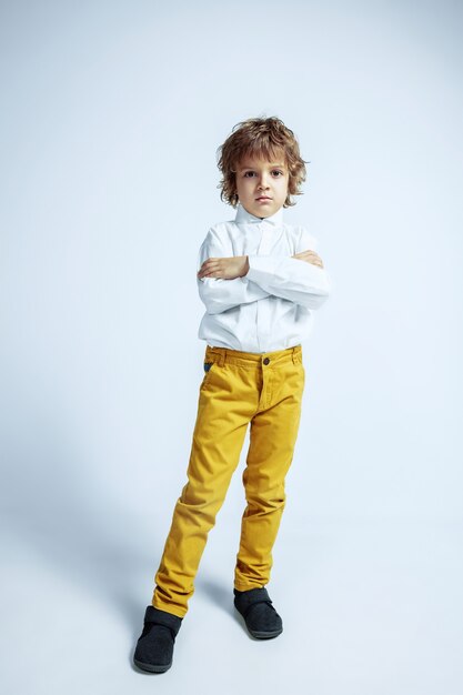 Pretty young boy in casual clothes on white wall. Fashionable posing. Caucasian male preschooler with bright facial emotions. Childhood, expression, having fun. Posing crossed hands.