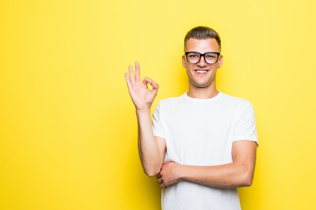 Pretty young boy shows okay sign dressed up in white t-shirt and transperent glasses isolated on yellow