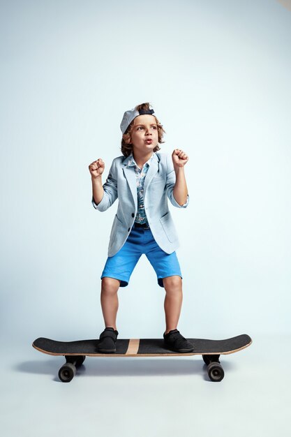 Pretty young boy on skateboard in casual clothes on white studio wall