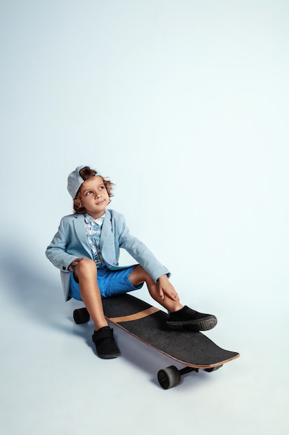 Pretty young boy on skateboard in casual clothes on white  wall. Riding and looks happy. Caucasian male preschooler with bright facial emotions. Childhood, expression, having fun.