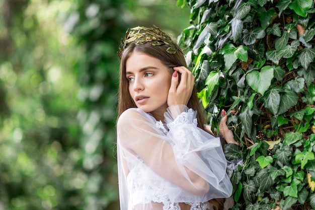 Pretty young bride in white lingerie and golden crown poses in a green garden