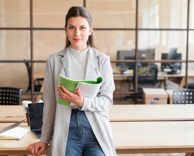 Pretty young businesswoman leaning on desk holding book looking at camera in office