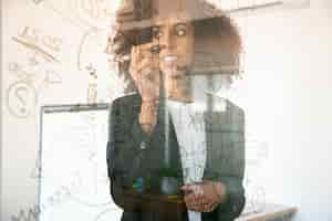 Free photo pretty young businesswomen writing on glass board. confident experienced african american female manager holding marker and smiling in office room. strategy, business and management concept